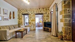 A cozy living room with a stone wall, a sofa, a coffee table, a TV, and a dining area in the background. - Apartments Panethimos (Platanias)