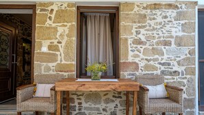 A stone wall with a wooden table and wicker chairs, a window with curtains, and a door with a metal gate. - Apartments Panethimos (Platanias)