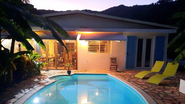 A swimming pool with a stone border, a covered patio area, and a house with a blue door and windows. - Ideally located, independent room with swimming pool (Le Marin)