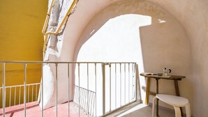 Ein Balkon mit weißer Reling, einem kleinen Holztisch und einem Stuhl, sowie eine gelbe Wand.