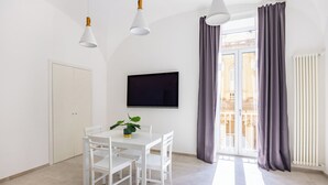 Ein modernes, helles Zimmer mit Esstisch, Stühlen, einer Topfpflanze, einem Flachbildfernseher und großen Fenstern mit Vorhängen.