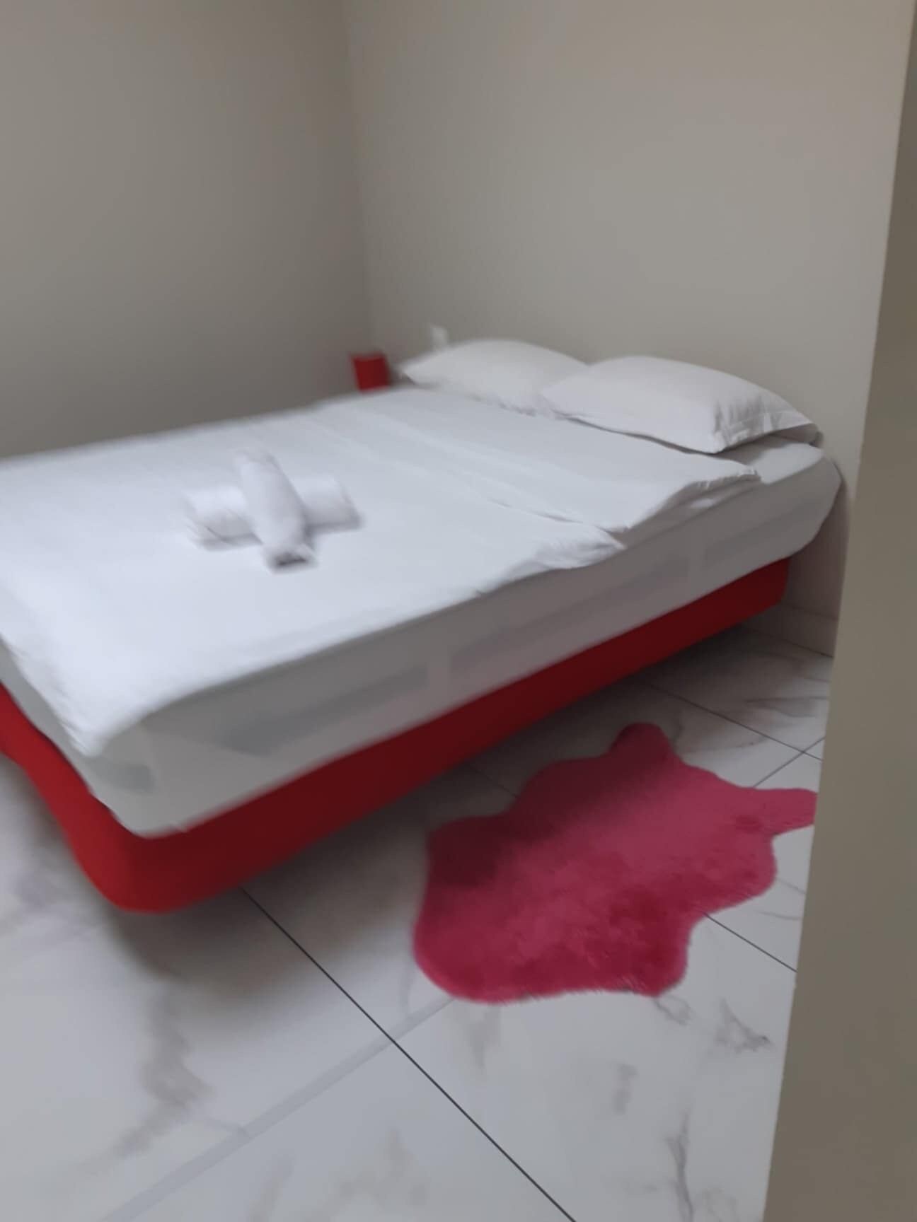 Ein kleines, ordentlich bezogenes Bett mit weißer Bettwäsche und einem roten Gestell, platziert in einem Zimmer mit weiß gefliestem Boden und einem rosa Teppich.