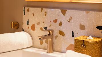 A bathroom with a white sink, a wooden-framed mirror, a terrazzo wall, a towel, and a small wicker box.