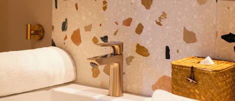 Un baño con un lavabo blanco, un espejo con marco de madera, una pared de terrazo, una toalla y una pequeña cesta de mimbre.