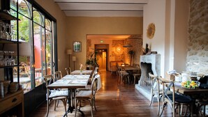 Un espace repas avec des tables et des chaises en bois, une cheminée en pierre et une vue dégagée grâce à de grandes fenêtres.