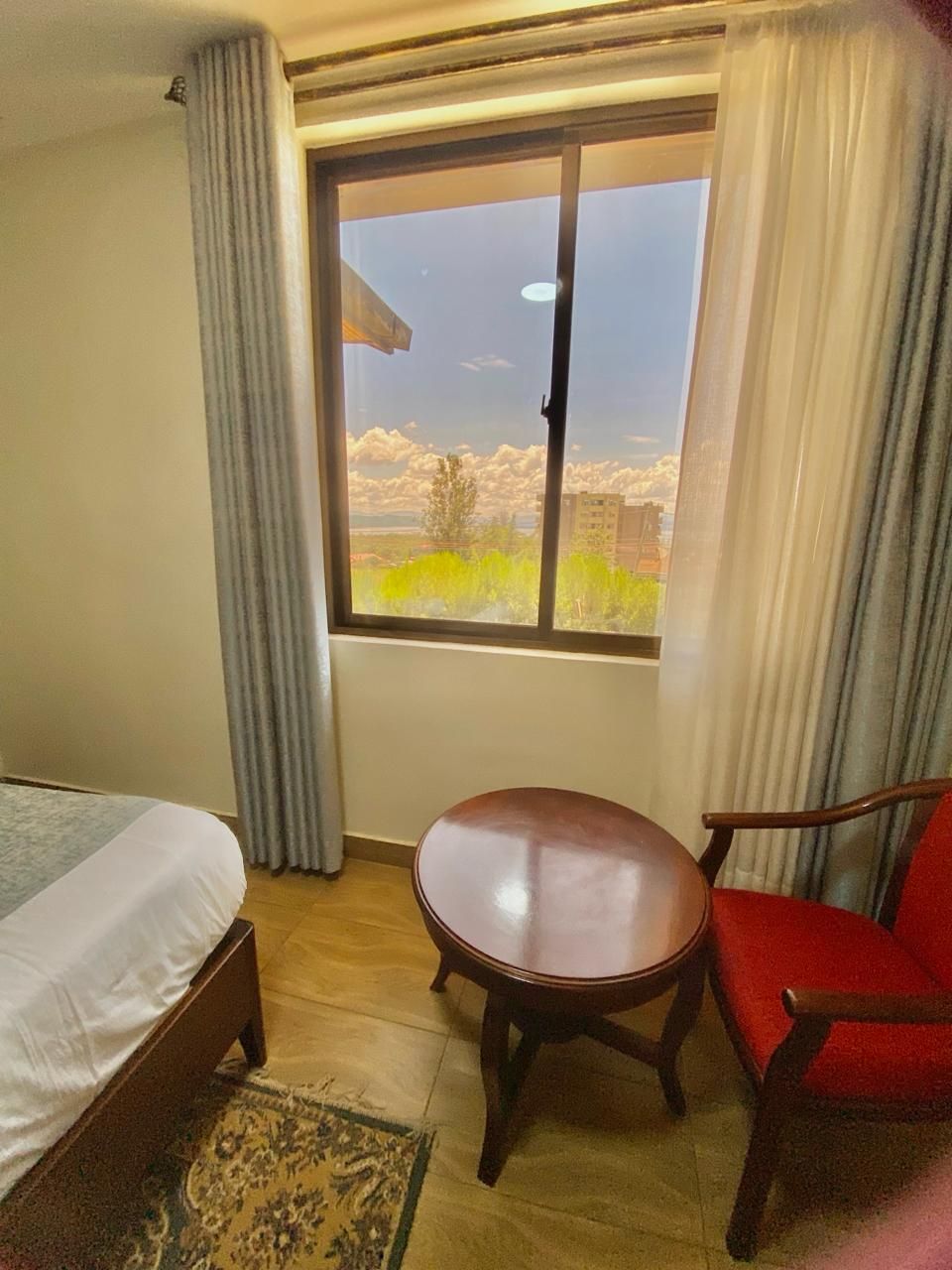 A hotel room with a bed, a small table, a red chair, and a window with a view of a building and trees.