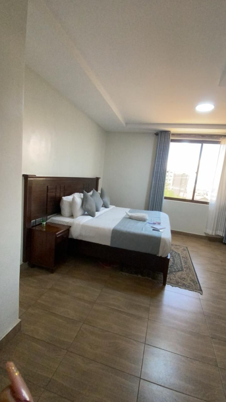 A hotel room with a bed, a nightstand, a window with curtains, and a view of buildings outside.