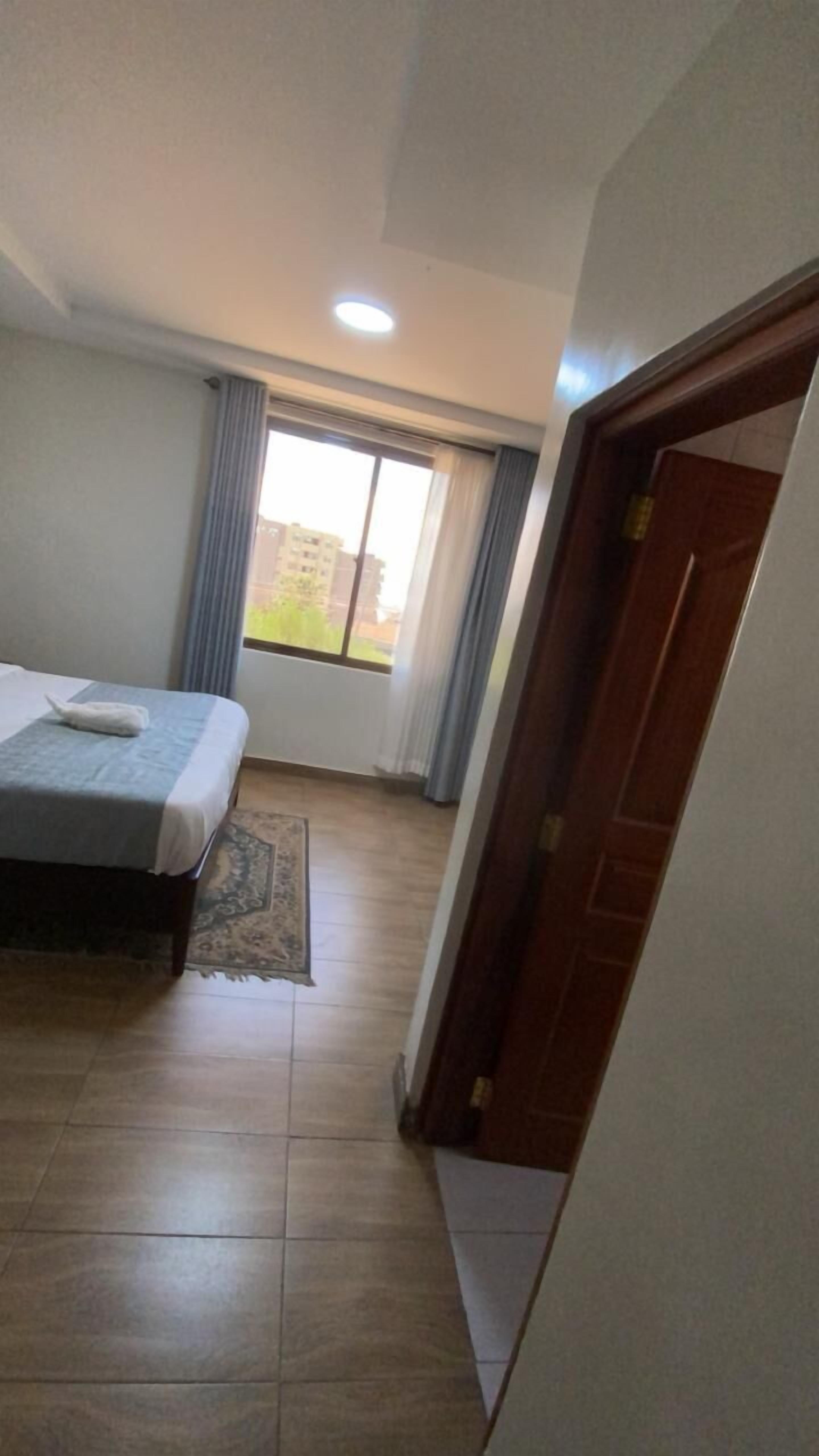 A hotel room with a bed, a window with curtains, a tiled floor, and a door.