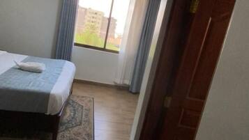 A hotel room with a bed, a window with curtains, a tiled floor, and a door.