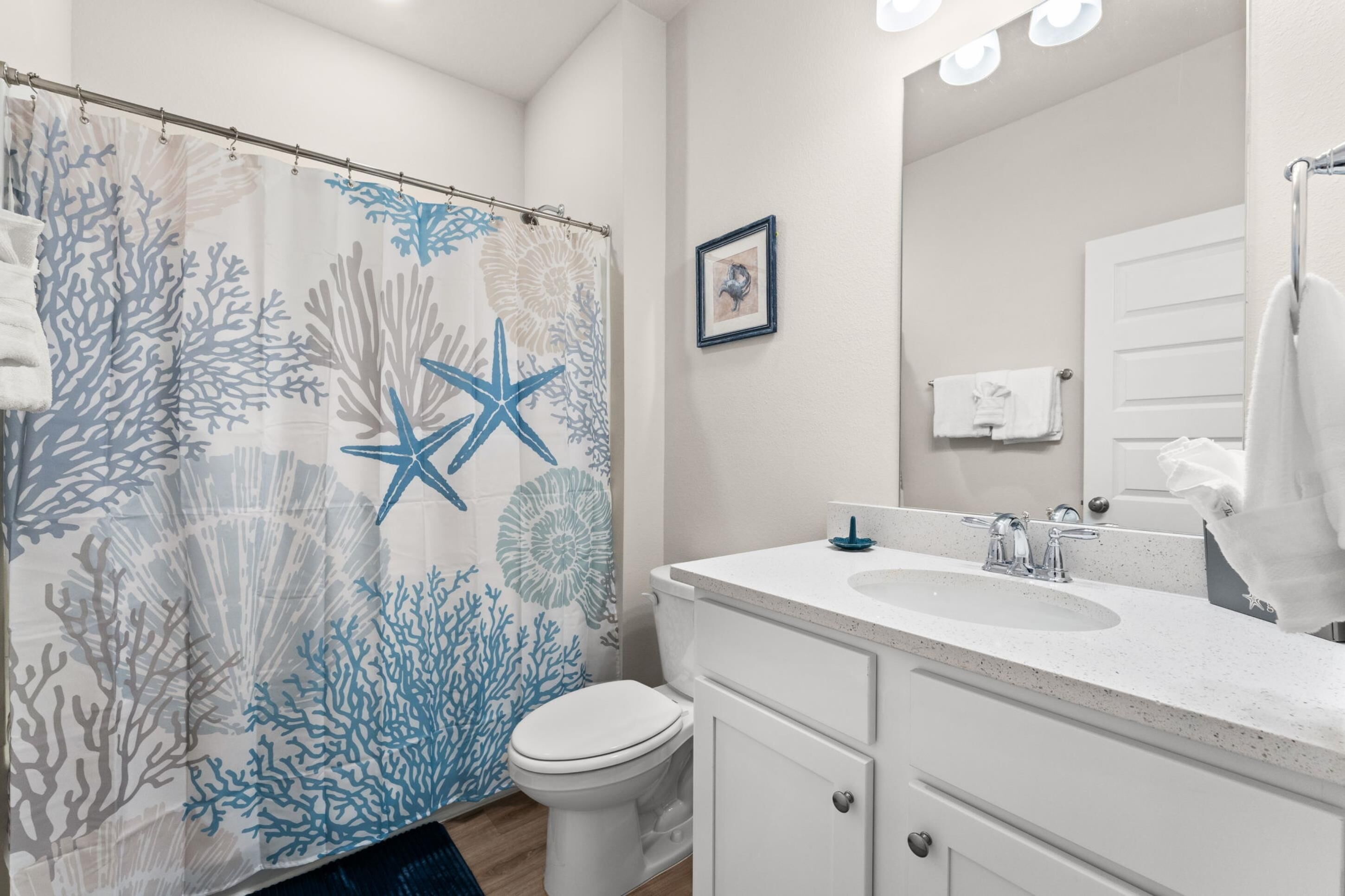 A bathroom with a shower curtain featuring a coral and starfish pattern, a white toilet, a sink with a mirror, and a towel rack.