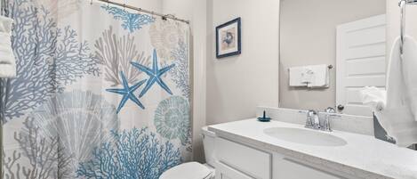 A bathroom with a shower curtain featuring a coral and starfish pattern, a white toilet, a sink with a mirror, and a towel rack.