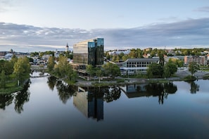 A modern building with a glass facade near a calm body of water, surrounded by greenery and other buildings.