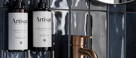 A bathroom with a white sink, two bottles of Artisan body lotion, and a copper faucet.