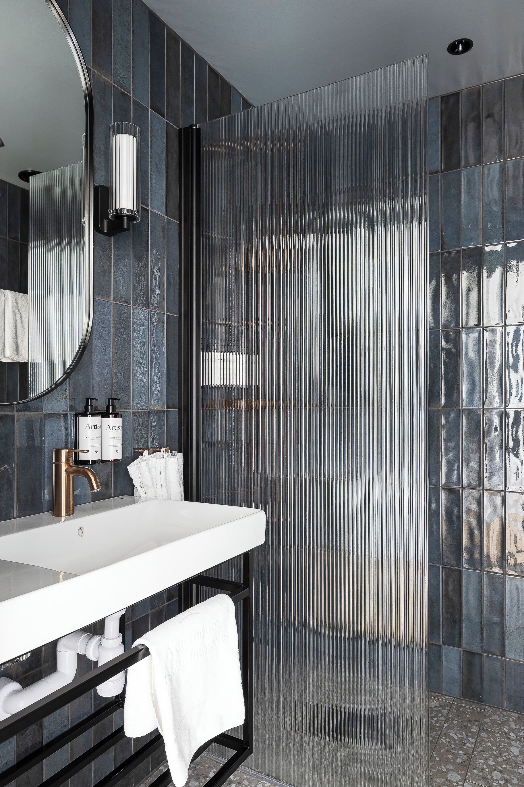 Un baño moderno con un lavabo blanco, un espejo grande y una zona de ducha con separador metálico acanalado.