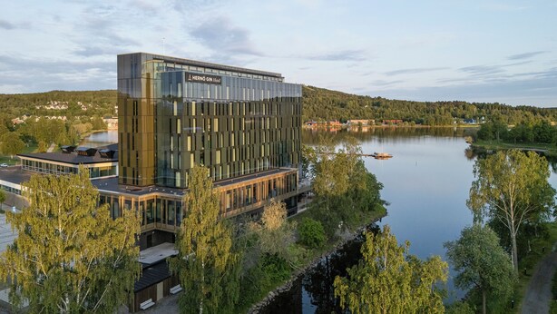 Un hôtel moderne à la façade en verre, situé au bord d’un lac paisible, entouré de verdure.