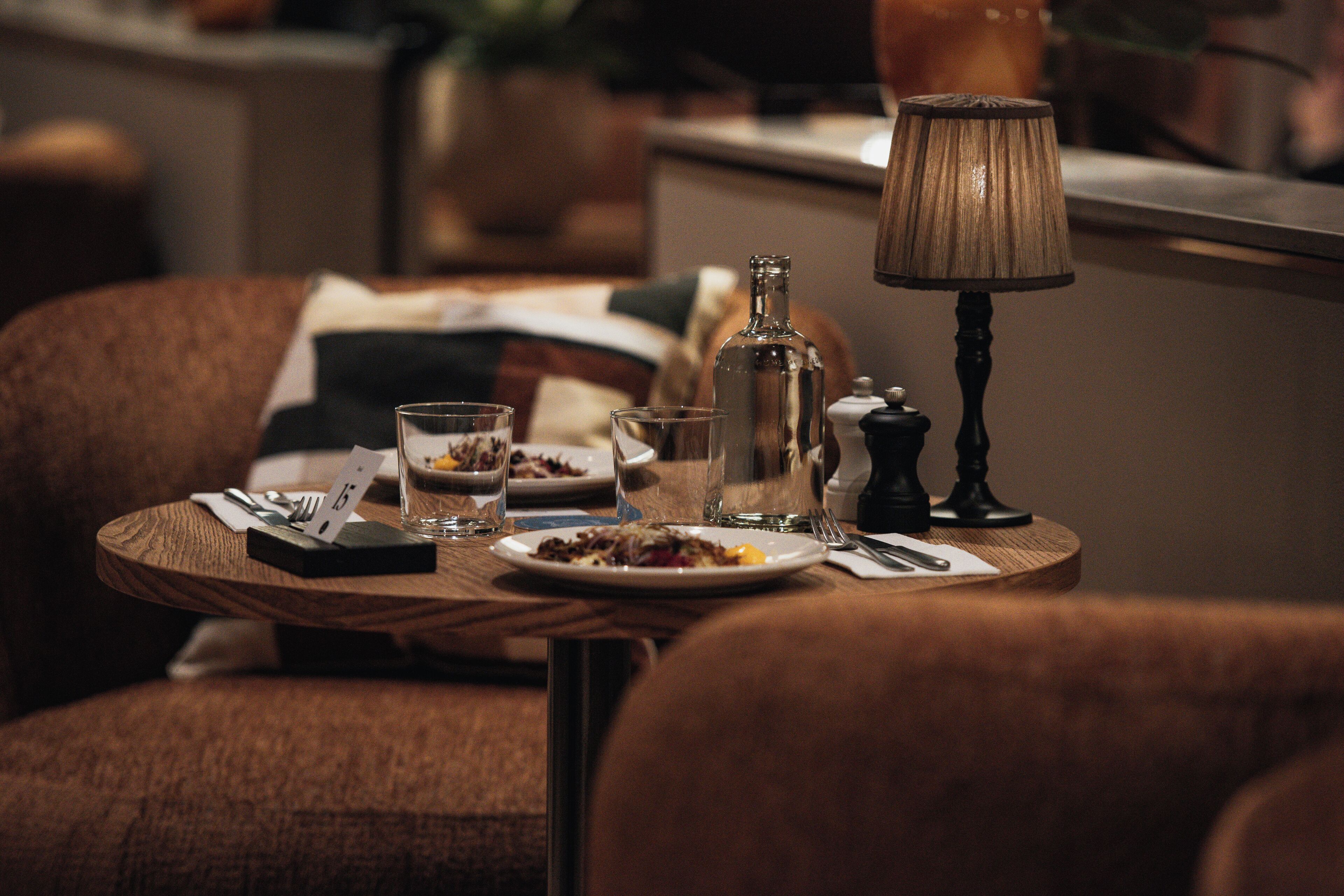 A dining table with plates, glasses, cutlery, and a lamp in a dimly lit room with upholstered chairs.