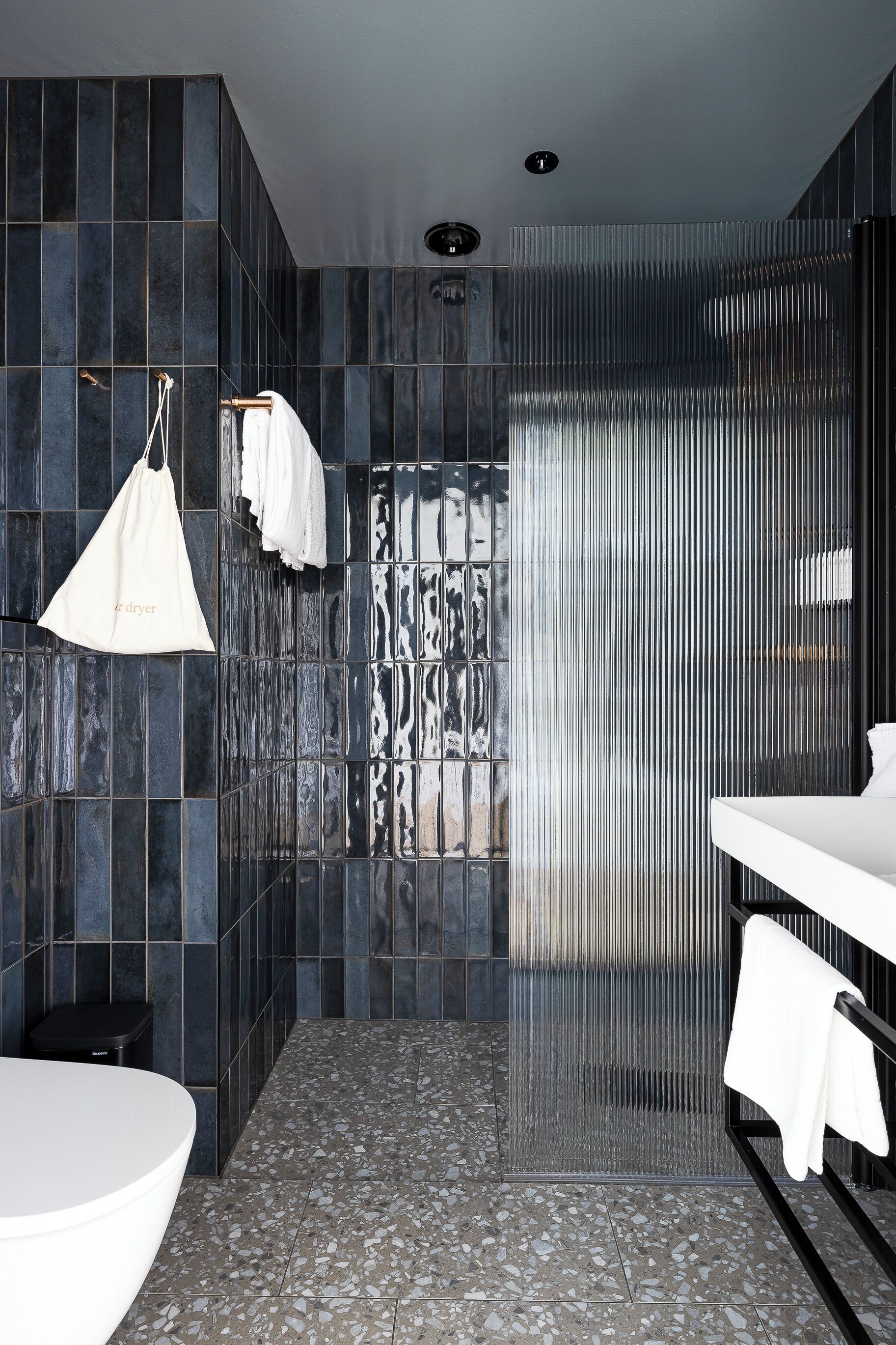 Un baño moderno con baldosas oscuras, una bañera blanca, ducha con puerta de cristal y un perchero para toallas.