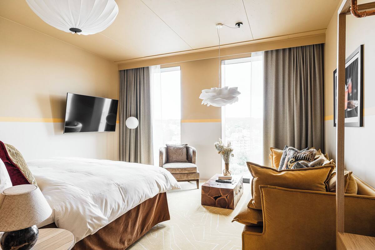 Une chambre d’hôtel moderne avec un grand lit, un canapé, un fauteuil, un téléviseur fixé au mur et de grandes fenêtres avec des rideaux.