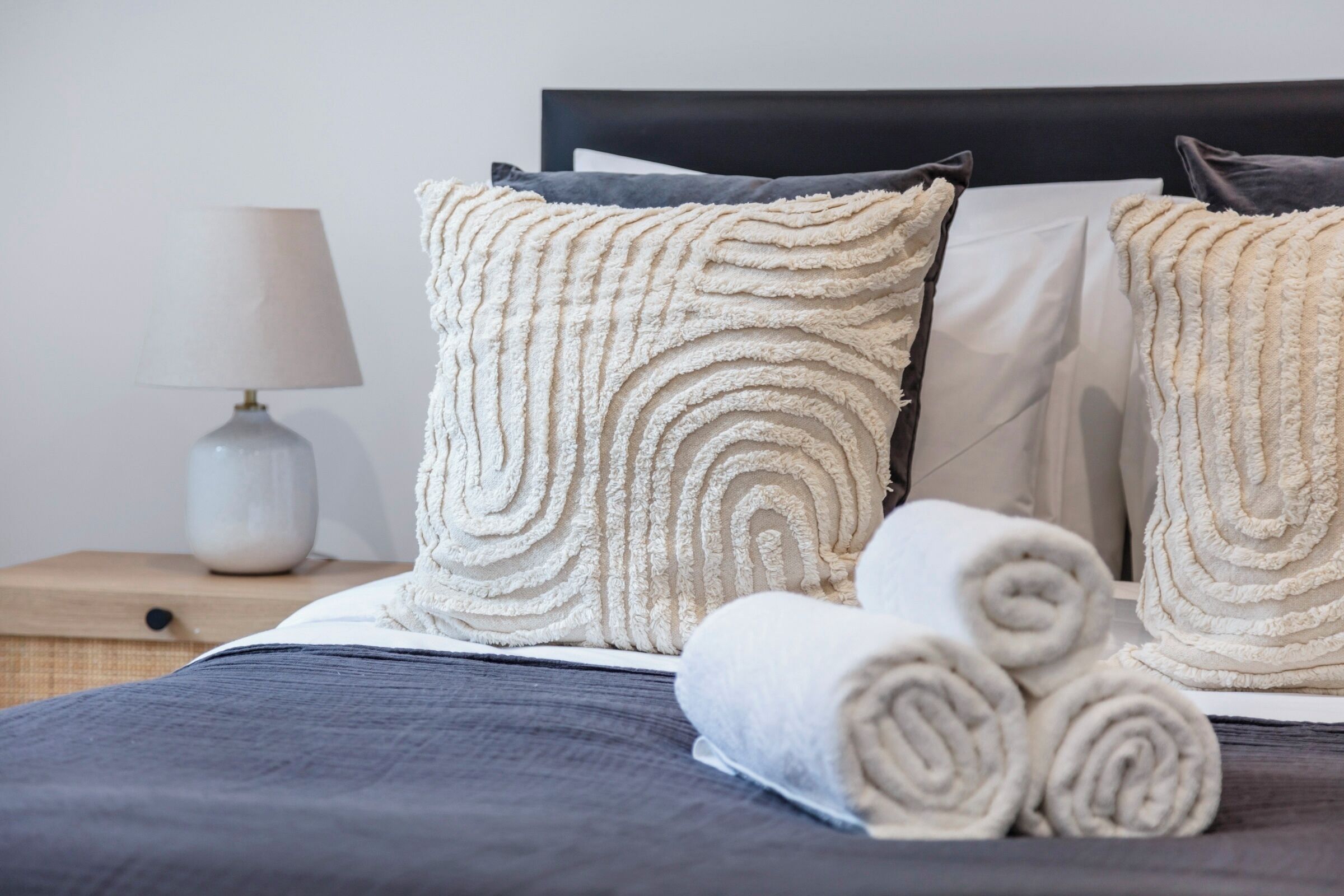 A neatly made bed with rolled towels, a lamp, and a nightstand with a vase.