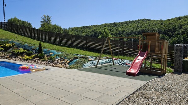 Piscina con tobogán, rodeada por una valla de madera y vista a una colina boscosa.