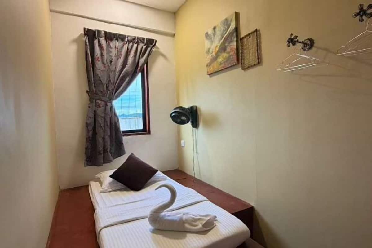 A small room with a bed, a window with curtains, a wall-mounted lamp, and a towel shaped like a swan on the bed.
