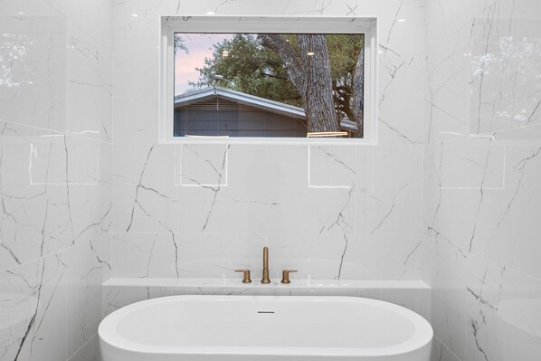 Ein modernes Badezimmer mit weißen Marmorwänden, einer großen Badewanne und einem Fenster mit Blick auf einen Baum und ein Gebäude.