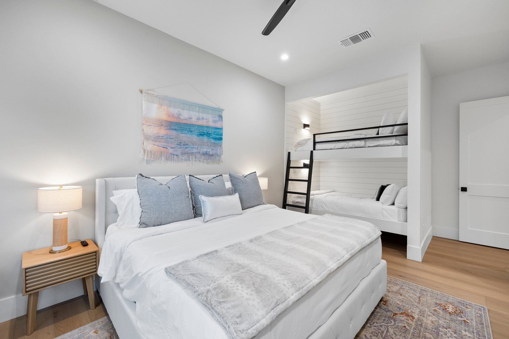 Un dormitorio moderno con una cama grande, una cama más pequeña, una mesita de noche con una lámpara, un estante en la pared y un cuadro de una playa.