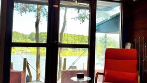 A room with large windows offering a view of a lake and trees. There is a red chair and a small table with a cup. - Stunning Waterfront Home with Private Dock - Wright on the Water! (Belle Haven)