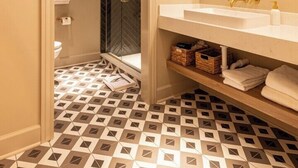 Une salle de bain avec un lavabo blanc, des robinetteries dorées, un miroir avec un cadre doré, des carreaux de sol à motifs et une porte donnant sur l’espace de la douche.