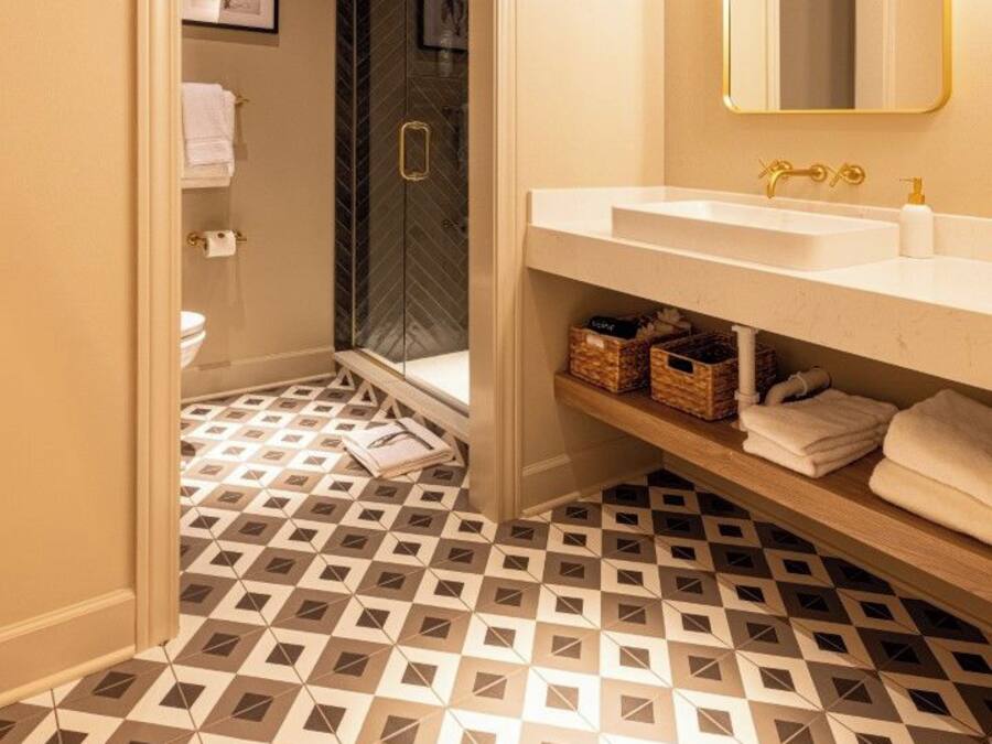 A bathroom with a white sink, gold fixtures, a mirror with a gold frame, patterned floor tiles, and a door leading to a shower area.