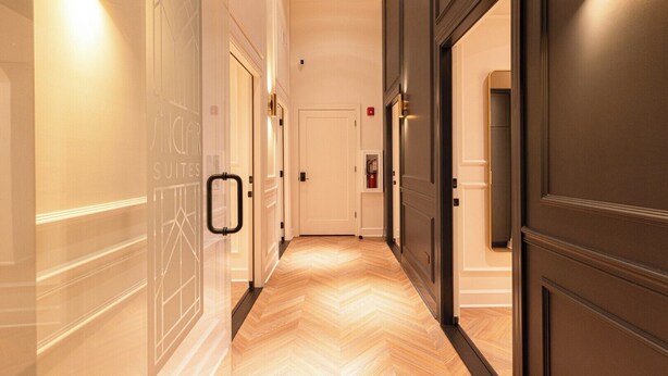Un couloir d’hôtel avec un parquet en chevrons, des murs blancs et des portes en bois foncé.
