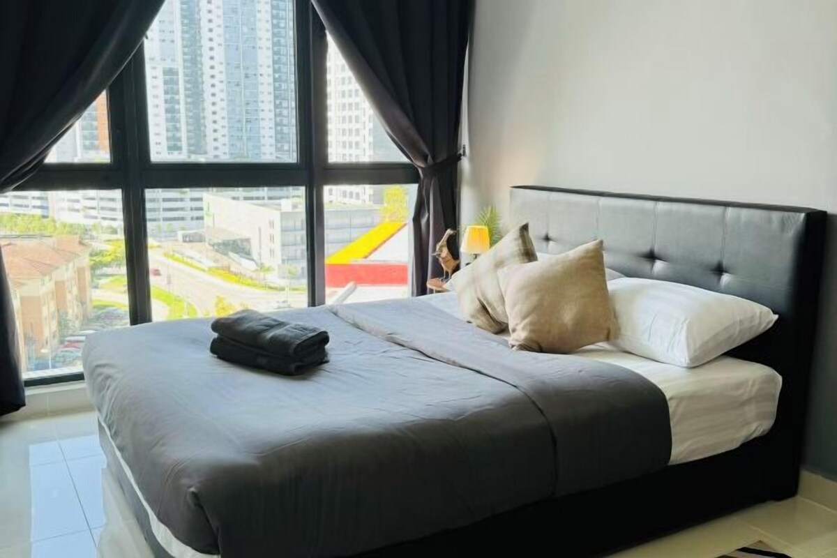 A bedroom with a large bed, a view of buildings through the window, and a lamp on the nightstand.