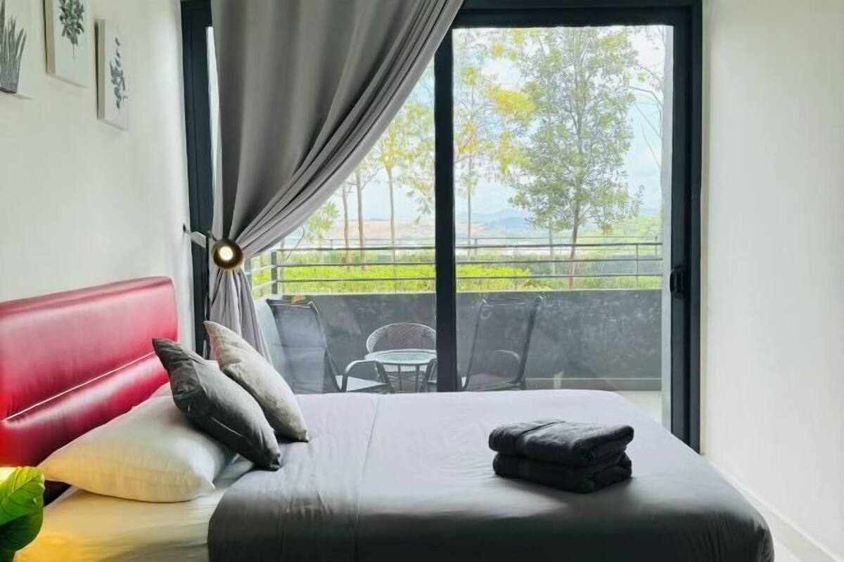 Une chambre d’hôtel avec un lit, une tête de lit rouge, un balcon avec des chaises et une vue sur la verdure et les montagnes.