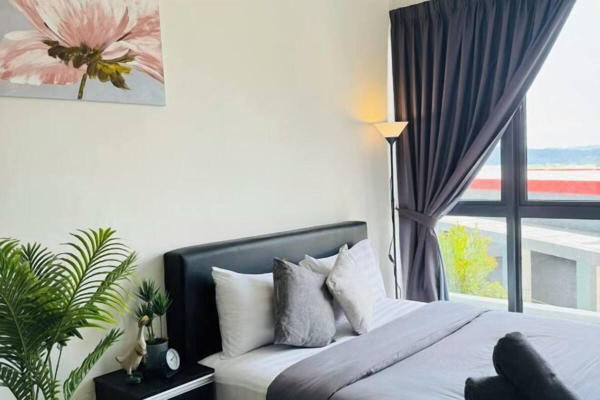 Une chambre à coucher avec un lit, des oreillers, une table de chevet, une lampe, une fenêtre avec des rideaux et une plante.
