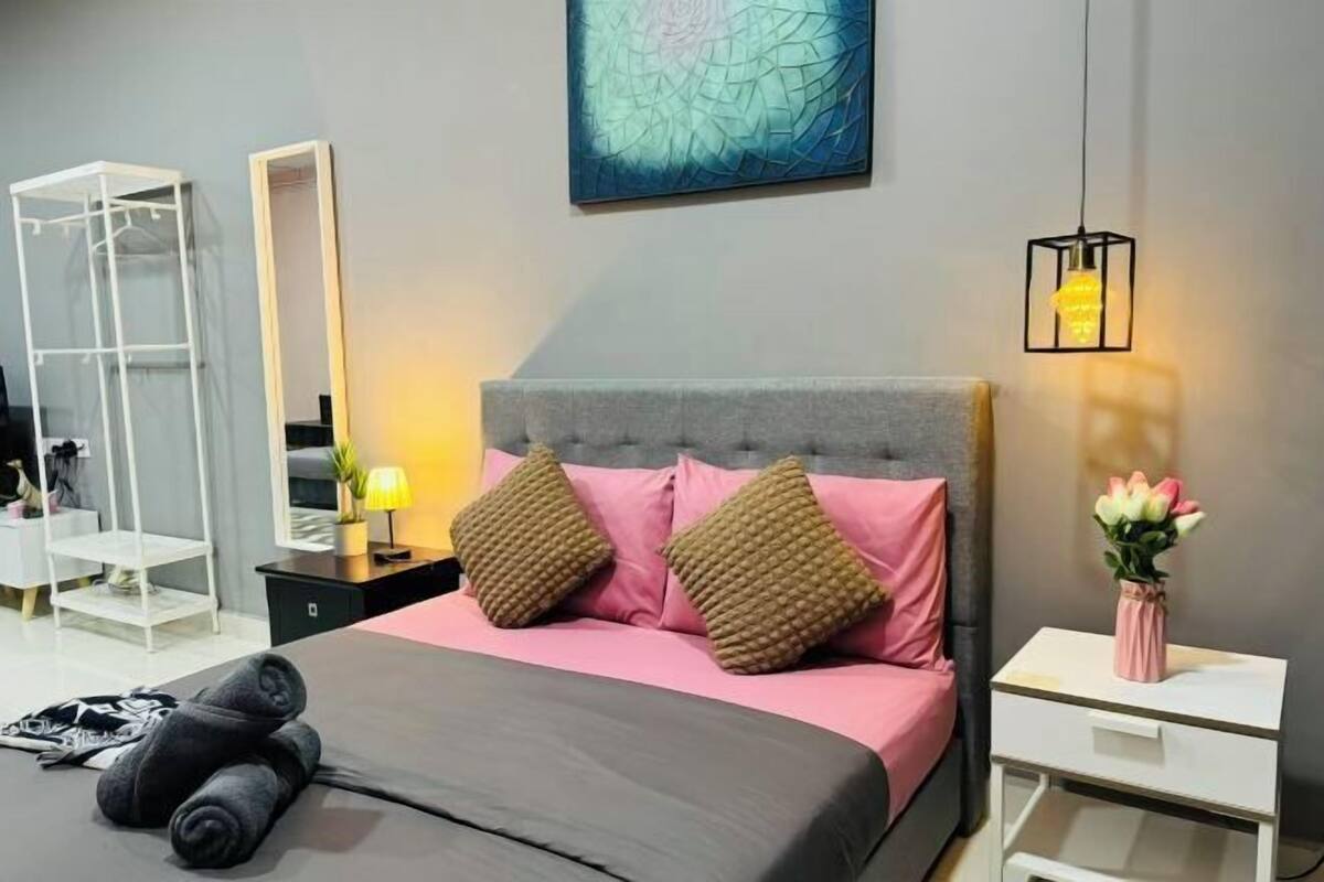 Une chambre moderne avec un lit gris, des coussins roses et bruns, une table de chevet avec une lampe, un miroir et une œuvre d’art murale.