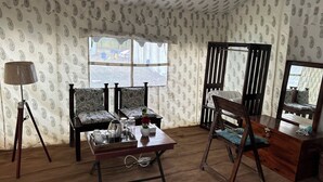 Ein Zimmer mit geprägtem Wandpapier, Holzboden, einer Sofagarnitur, einem Tisch mit Tablett und Gebäck, einer Lampe und einem Spiegel.