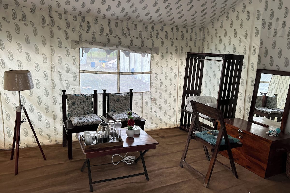 Quarto com papel de parede estampado, piso de madeira, sofá, mesa com bandeja de objetos, abajur e espelho.