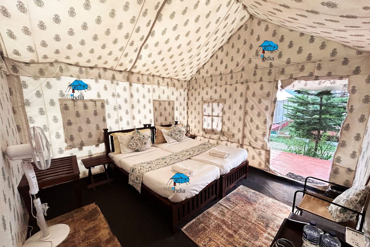 Quarto tipo tenda com cama, ventoinha, mesa pequena e janela com vista para o verde lá fora.