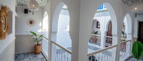 A corridor with white walls, patterned floor tiles, and decorative ceiling designs. There are potted plants and a hanging light fixture.