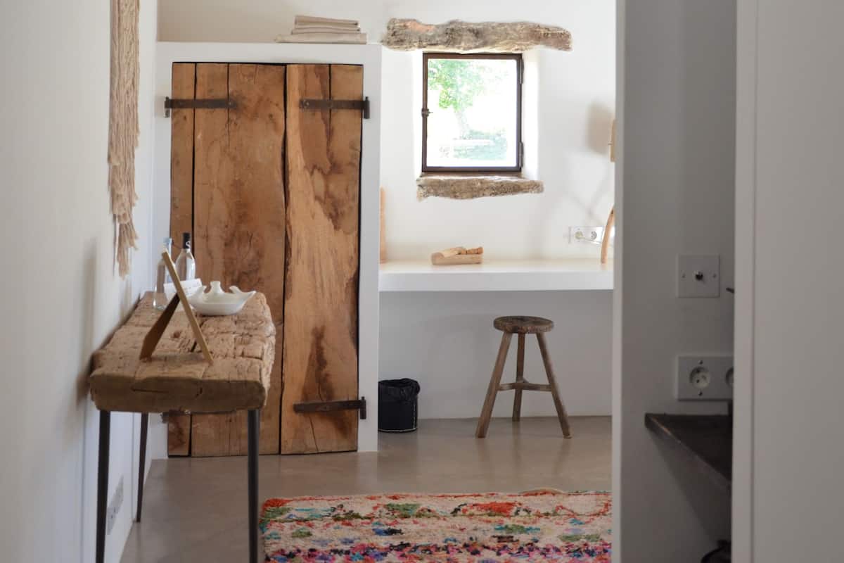 Een rustiek interieur met een houten plafond, een eenvoudige houten bank, een kleurrijk vloerkleed en een klein krukje.