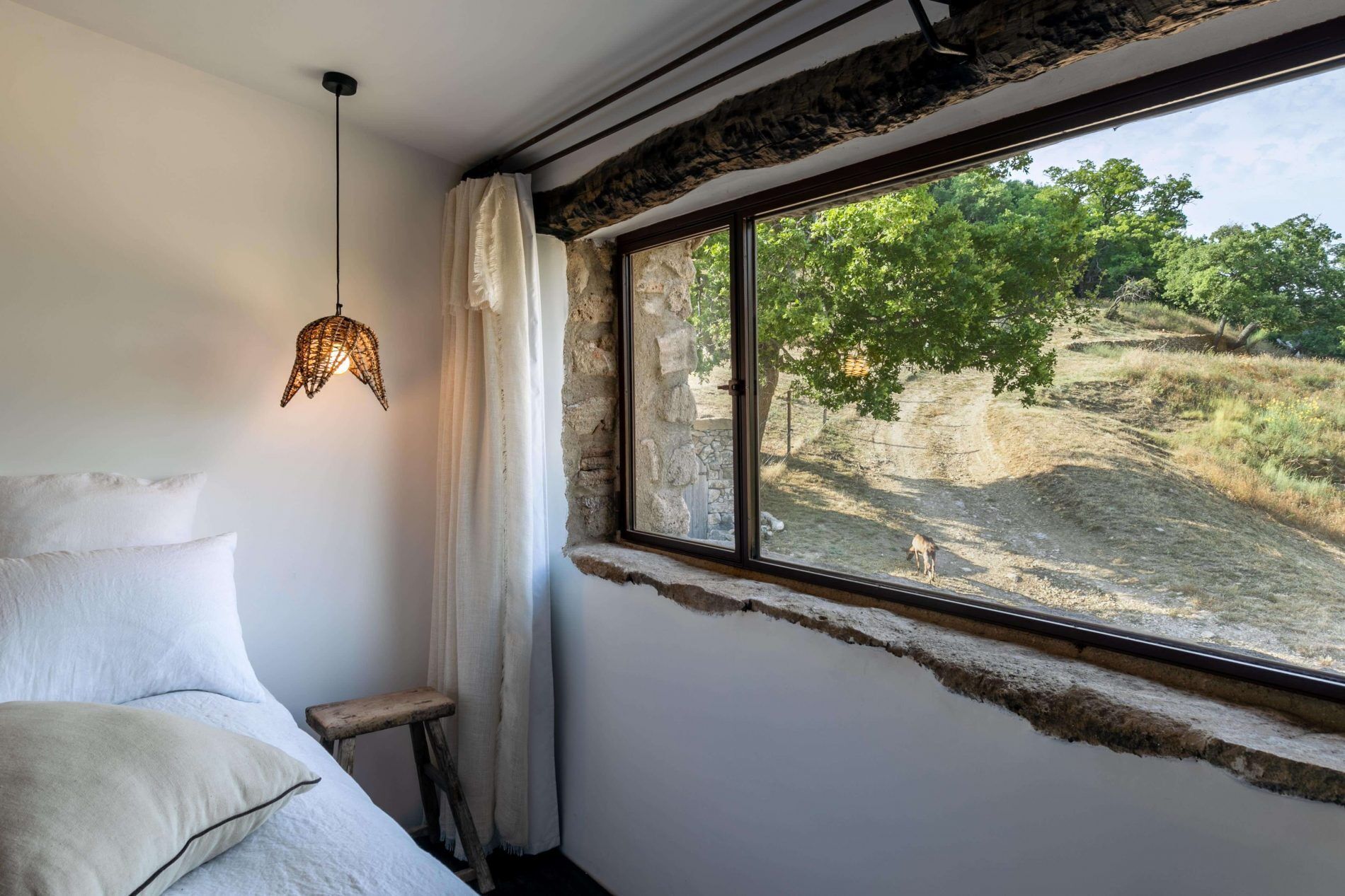 A room with a stone wall, a window with a view of a forest, a hanging light fixture, and a bed with pillows.