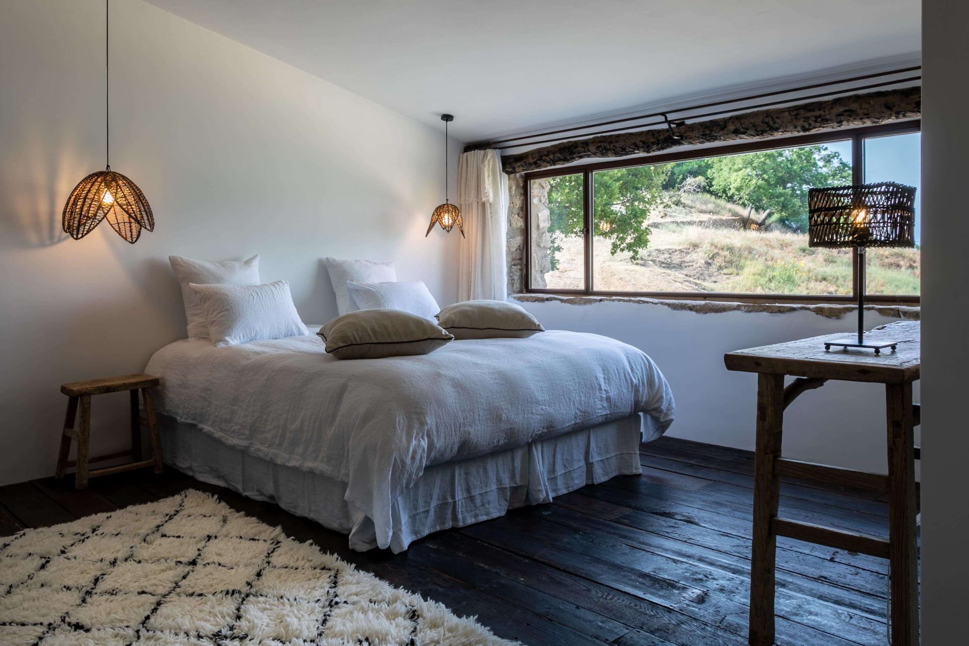 Een slaapkamer met een bed, twee bedlampen, een houten nachtkastje, een raam met uitzicht op bomen en een zacht, wit vloerkleed.