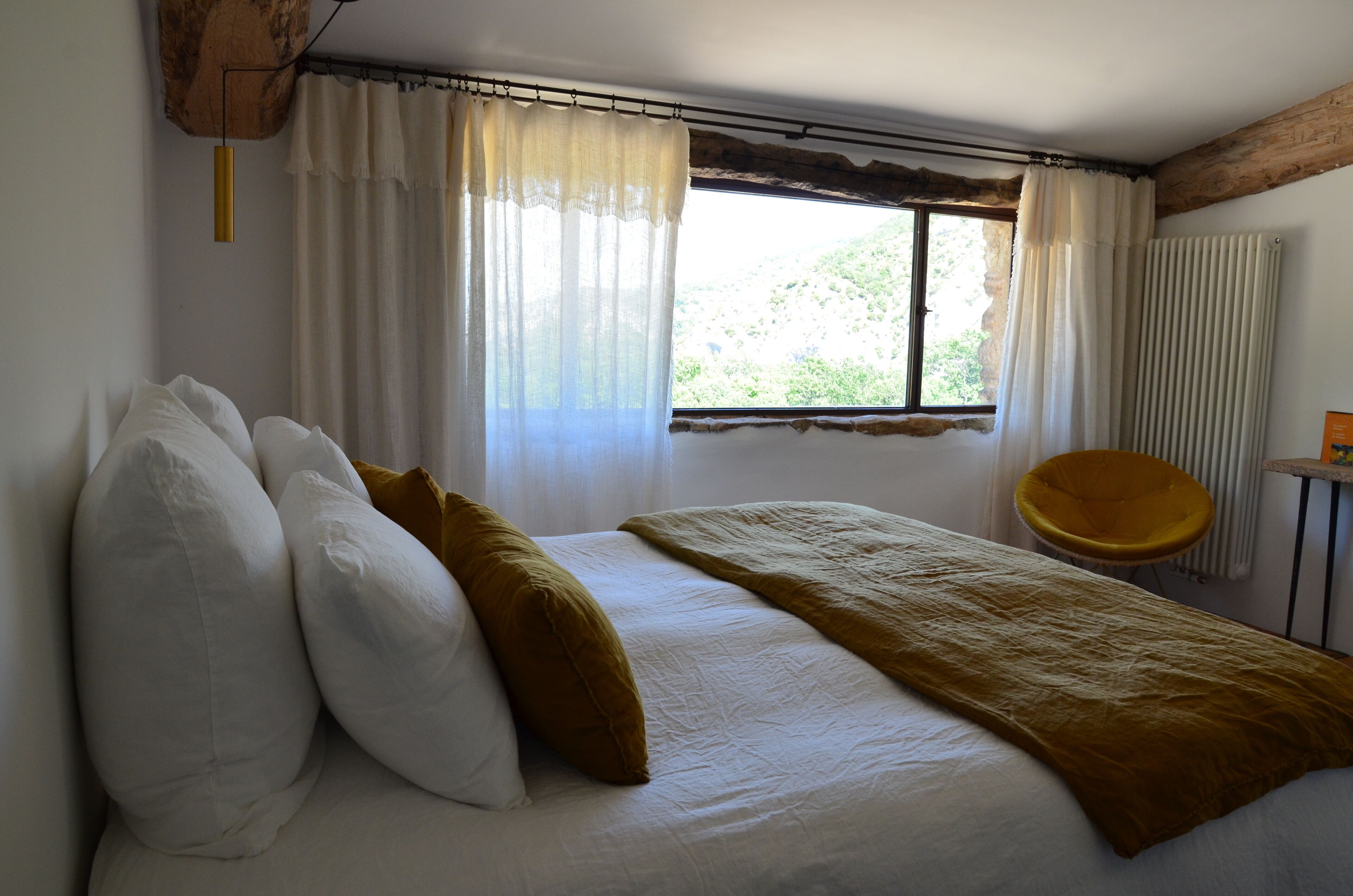 A bedroom with a bed, white and mustard pillows, a blanket, a chair, and a window with curtains.