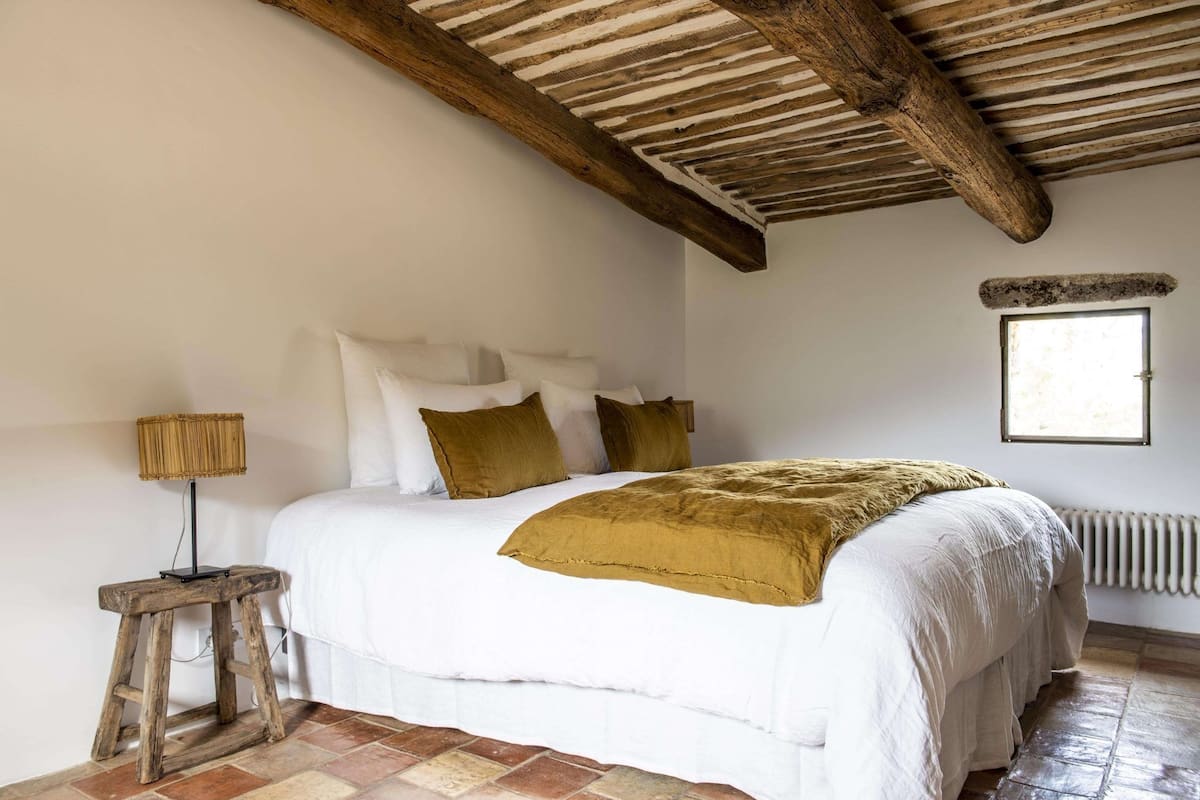 Een slaapkamer met een houten plafond, een bed met wit en bruin beddengoed, een kleine tafel met een lamp en een raam.