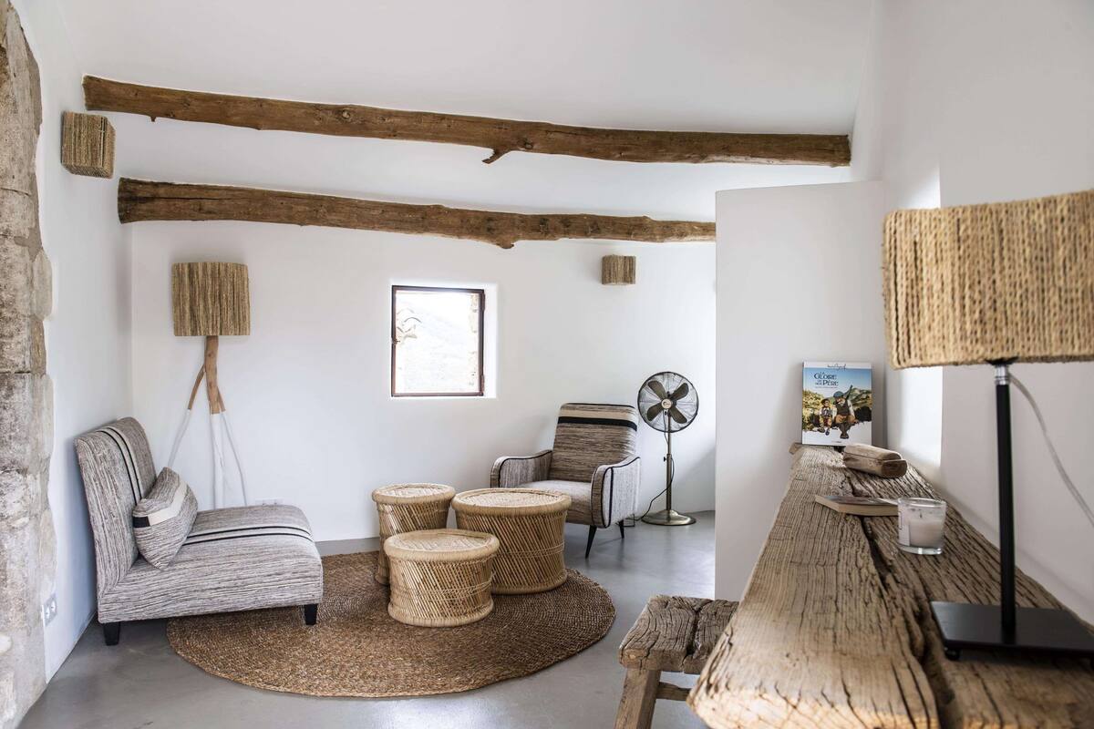 Een gezellige kamer met een rustiek houten plafond, een geweven vloermat, een bank, een klein rond tafeltje, een ventilatorkachel en een lamp.