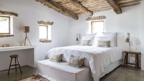 Une chambre avec un grand lit, un petit tabouret, un banc et une fenêtre.