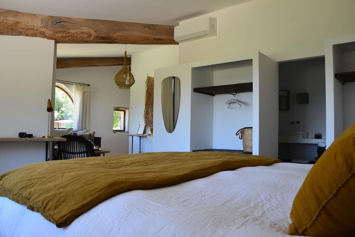 Een slaapkamer met een groot bed, een bureau en een stoel. Er is een raam met gordijnen, een hanglamp en een spiegel aan de muur.