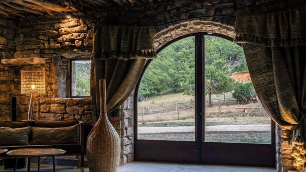 Une pièce rustique aux murs de pierre, dotée d’une grande fenêtre en arc de cercle, d’un panier en osier et d’une table.