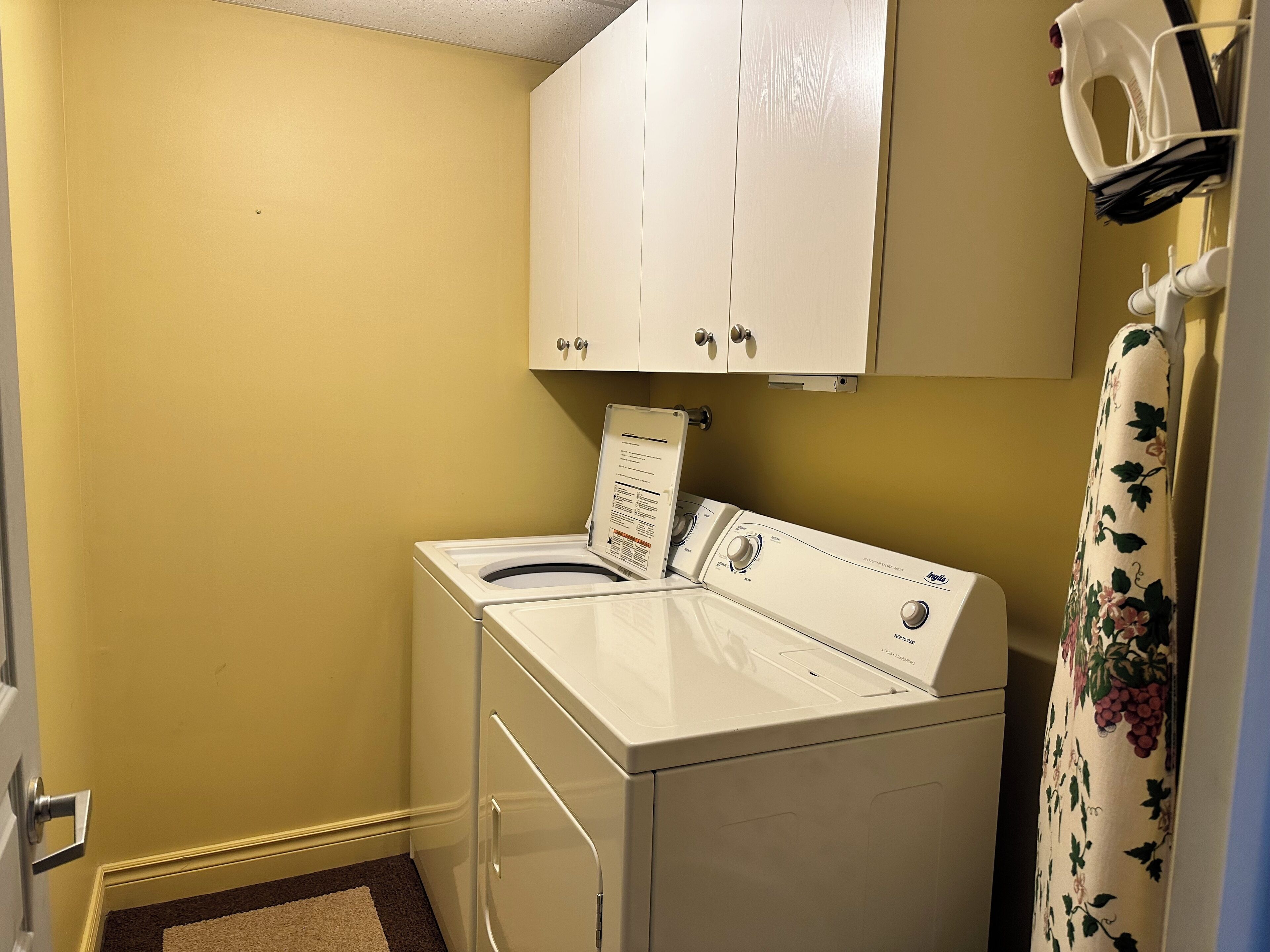 A compact laundry room with a front-loading washer and dryer, a white cabinet above, and a floral curtain on the right.