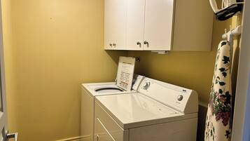 A compact laundry room with a front-loading washer and dryer, a white cabinet above, and a floral curtain on the right.